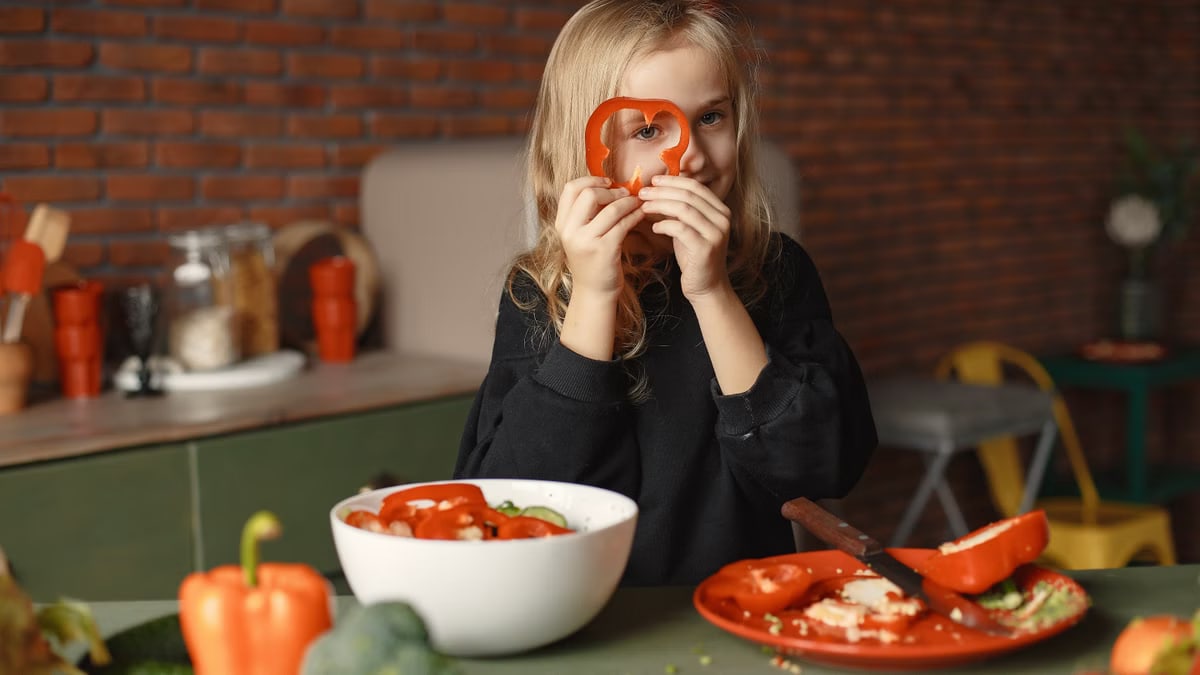 Funny cheerful girl looking through slice of pepper while preparing healthy vegetarian salad at table with plate with knife and salad bowl